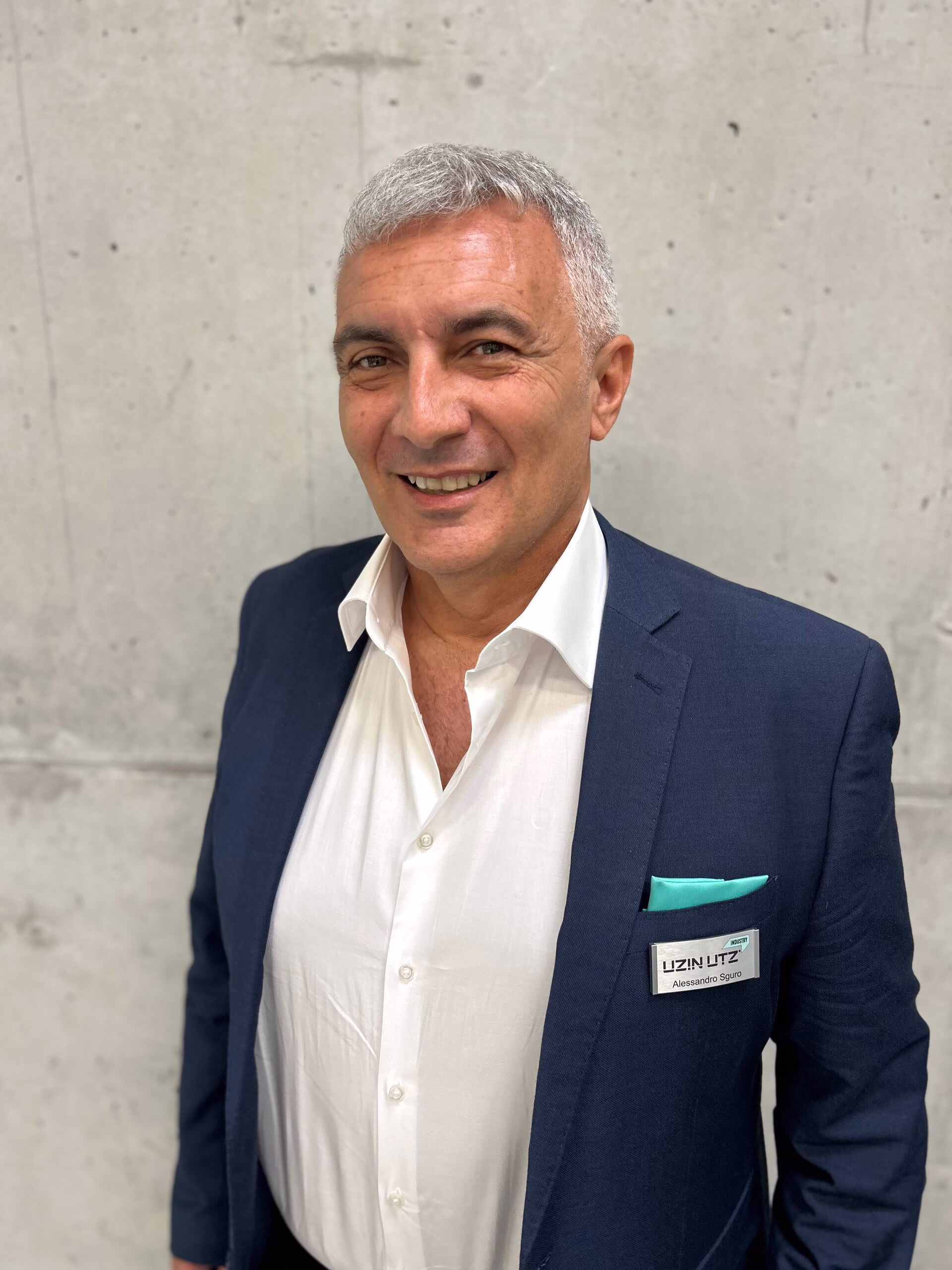 Ansprechpartner Italien; Ein Mann mit kurzen grauen Haaren, der einen marineblauen Anzug und ein weißes Hemd trägt, steht vor einer schlichten, hellgrauen Betonwand und lächelt. An seinem Anzug trägt er ein mintfarbenes Einstecktuch und ein Namensschild.