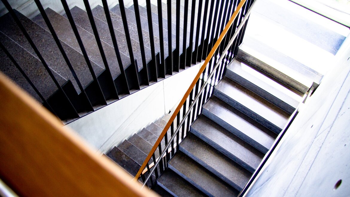 Eine moderne Treppe mit schwarzen Geländern und Holzhandläufen, von oben in einem Winkel aufgenommen, wobei die Stufen und Schatten ein geometrisches Muster bilden.