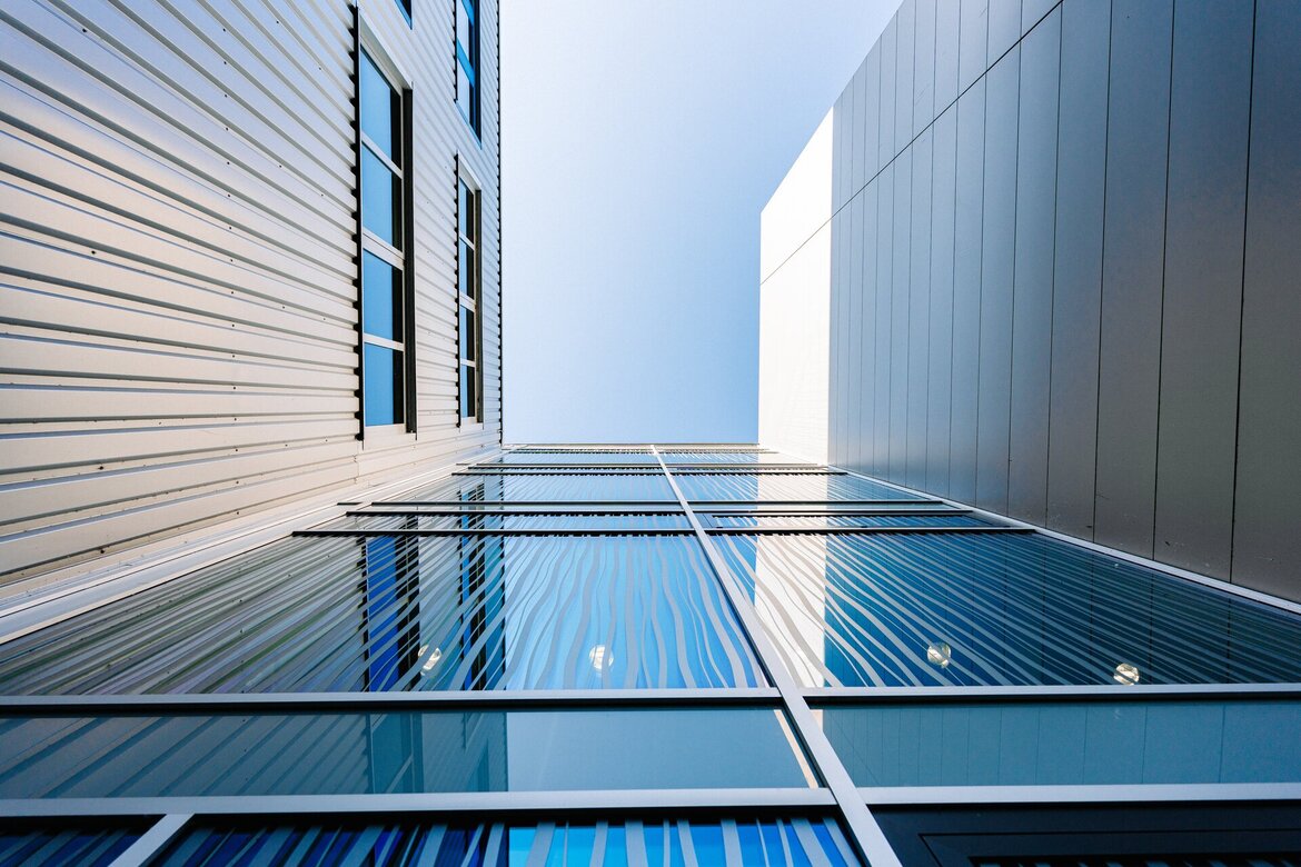 Eine Aufnahme aus tiefem Blickwinkel, die zu modernen Gebäuden mit Metallverkleidung und Glasfenstern hinaufführt, in denen sich der blaue Himmel spiegelt. Die Perspektive erzeugt starke vertikale und diagonale Linien, die die Höhe der Gebäude und ihre geometrischen Formen betonen.