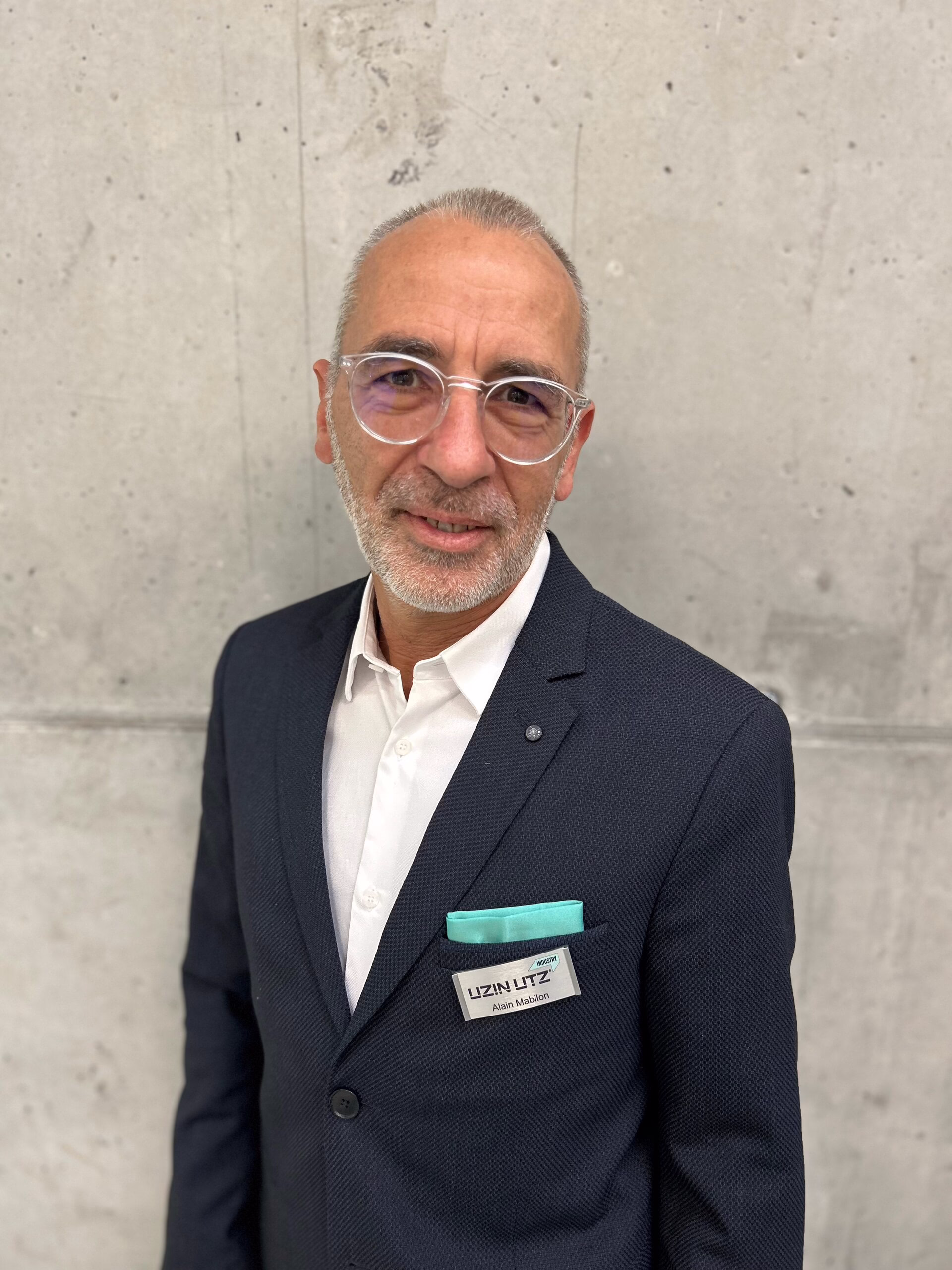 Ansprechpartner Frankreich; Ein Mann mit Brille und kurzem Bart steht vor einer grauen Betonwand und trägt einen dunklen Anzug, ein weißes Hemd sowie ein Namensschild mit der Aufschrift.