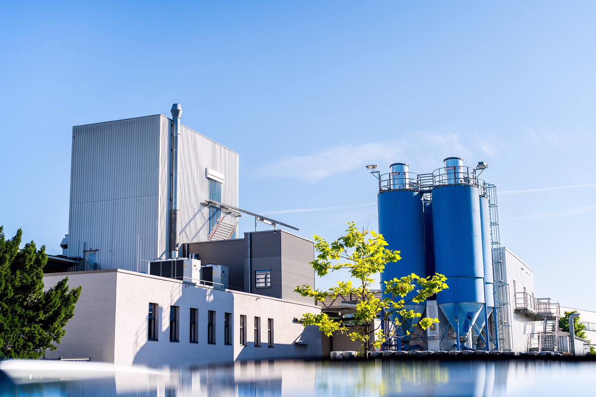 Moderne Industrieanlage mit einem weißen Gebäude, hohen blauen Silos und Metallkonstruktionen vor einem strahlend blauen Himmel. Im Vordergrund sind grüne Äste zu sehen.