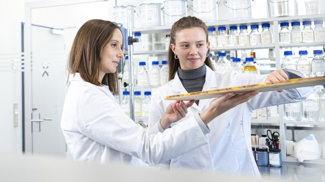 Zwei Wissenschaftler in Laborkitteln stehen in einem Labor und besprechen Informationen auf einem Klemmbrett. Im Hintergrund sind Regale mit Behältern und Laborgeräten zu sehen; die Szene wird durch ein Regal hindurch gezeigt.