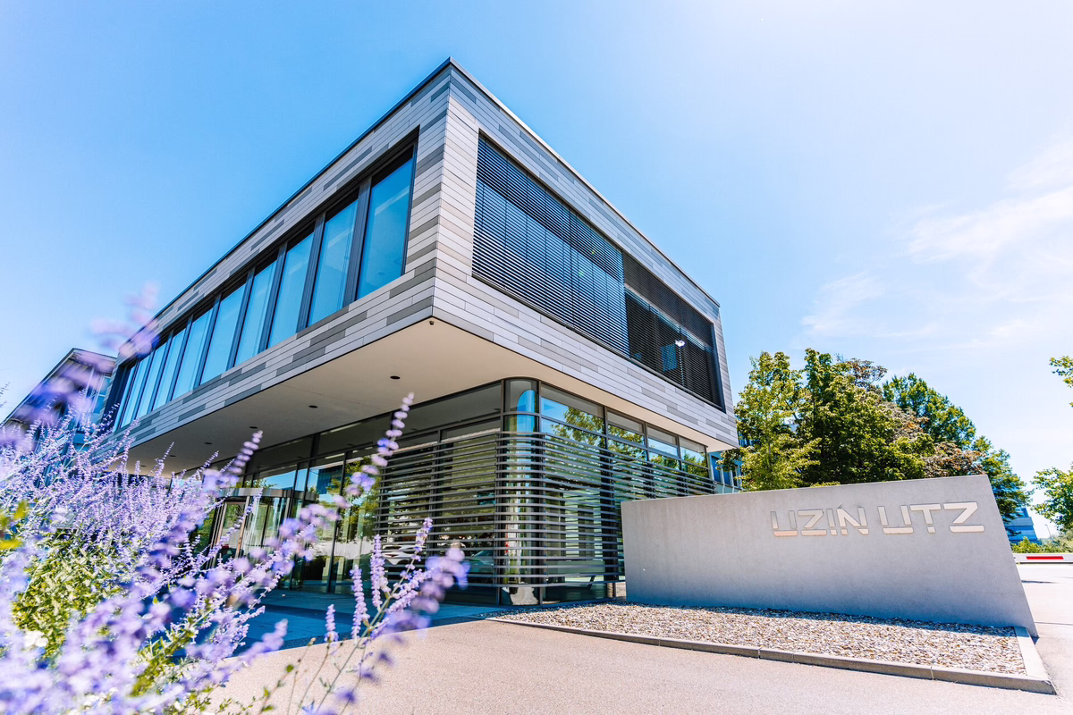 Modernes Bürogebäude aus Glas mit einem Schild mit der Aufschrift „UZIN UTZ“ vor dem Gebäude, umgeben von Grünanlagen und violetten Blumen, unter einem strahlend blauen Himmel.