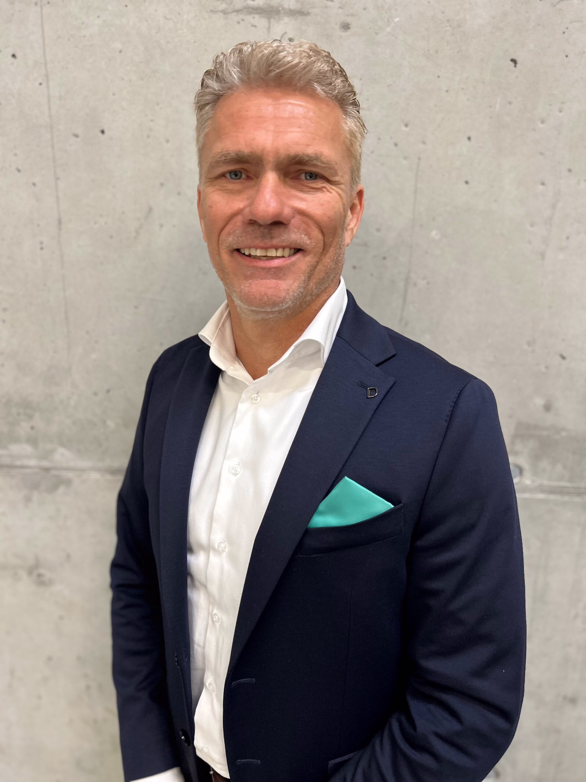 Ansprechpartner Niederlande; Ein Mann in einem marineblauen Anzug und einem weißen Hemd steht lächelnd vor einer grauen Betonwand, die linke Hand in der Hosentasche, aus der ein mintfarbenes Einstecktuch herausschaut.