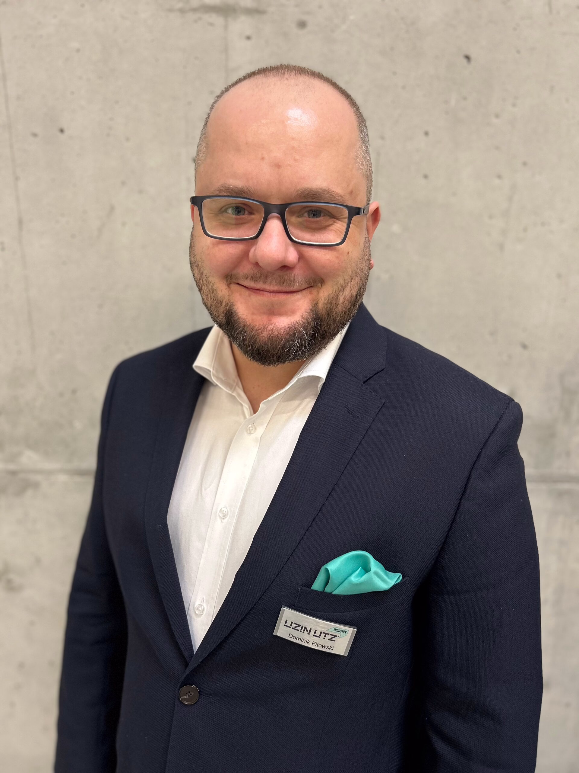 Ansprechpartner Polen; Ein Mann mit Brille und Bart, der einen dunklen Anzug, ein weißes Hemd und ein mintfarbenes Einstecktuch trägt, steht vor einer grauen Betonwand. An seiner Jacke befindet sich ein Namensschild, und er lächelt in die Kamera.